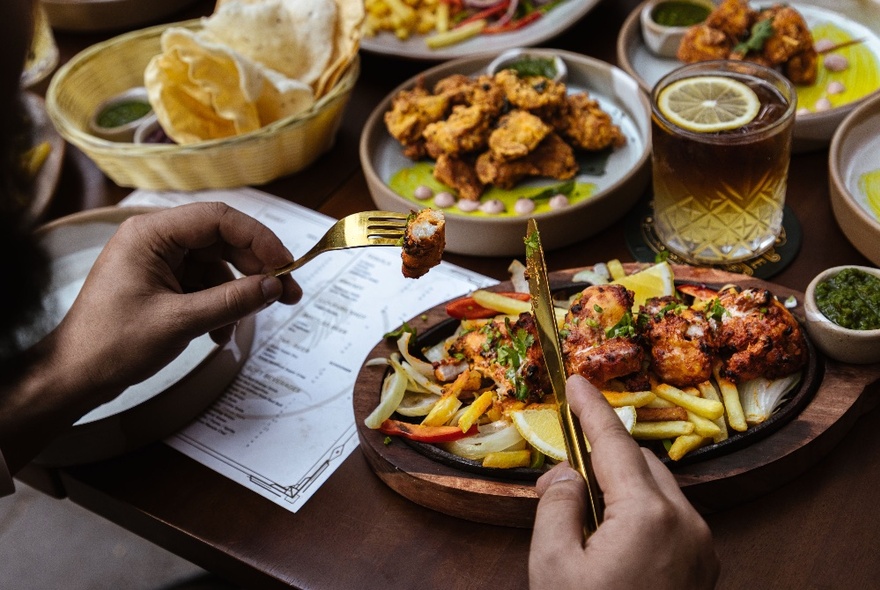 Looking at a table with various plates of Indian food and a diner with their cutlery poised to eat. 