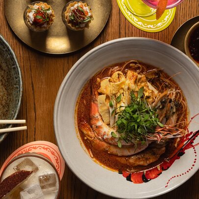 A chef's specialty laksa dish with prawns, noodles and a green garnish, alongside other malaysian dishes.