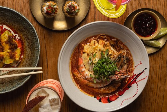 A chef's specialty laksa dish with prawns, noodles and a green garnish, alongside other malaysian dishes.