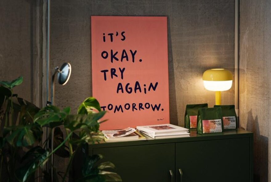 A pink sign that says 'It's okay. Try again tomorrow.' resting on a cabinet against a concrete wall.