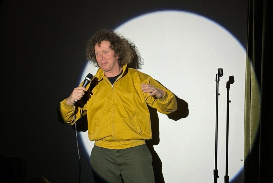A stand up comedian on a stage, holding a microphone, performing in a spotlight.