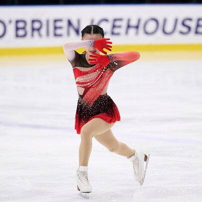 A figure skater wearing a red and black costume skating on an ice rink with the words O'BRIEN ICEHOUSE visible on a hoarding in the background.
