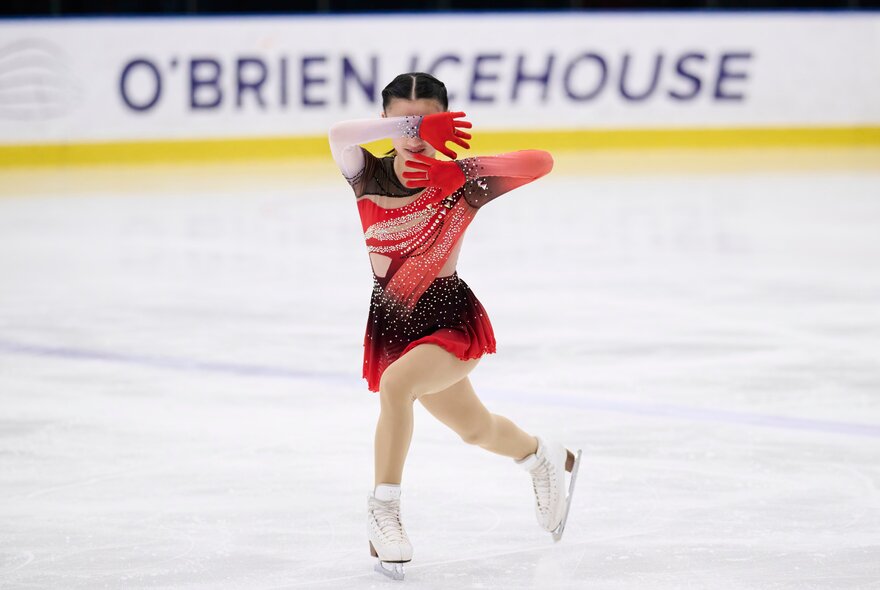 A figure skater wearing a red and black costume skating on an ice rink with the words O'BRIEN ICEHOUSE visible on a hoarding in the background.