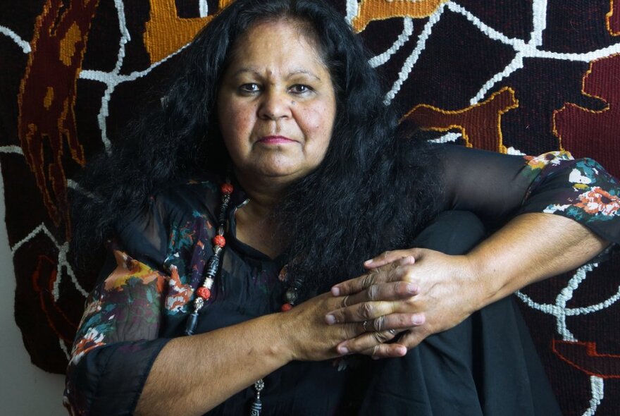 Artist Fiona Clarke seated in front one of her artworks, staring at the camera, with her fingers interlaced in front of her.