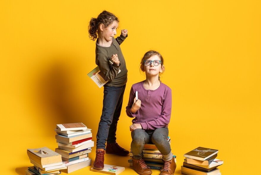 Two girls in a yellow room, one standing and one seated, next to piles of books