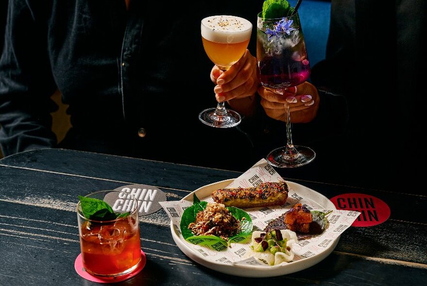 People clinking cocktails over a plate of small bites in a restaurant.