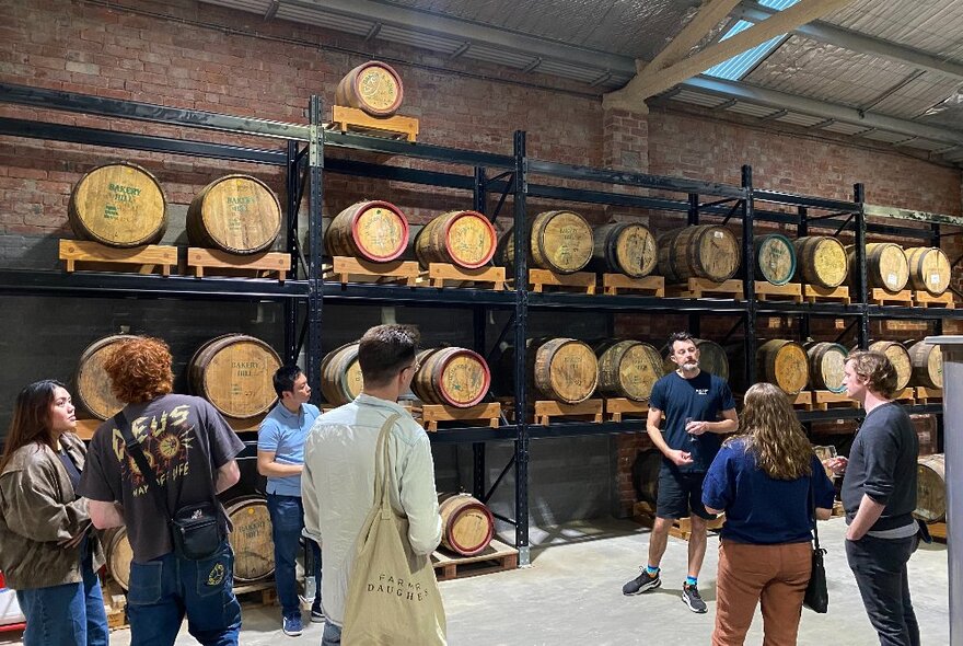 People standing in a distilling area with oak barrels stacked on shelves.