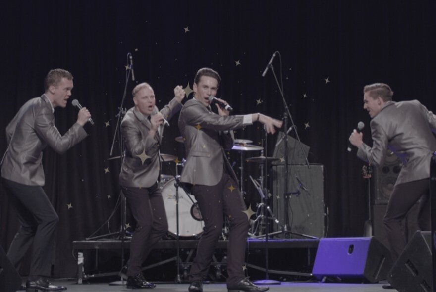 Vocal group performing on stage wearing matching shiny jackets and holding microphones.