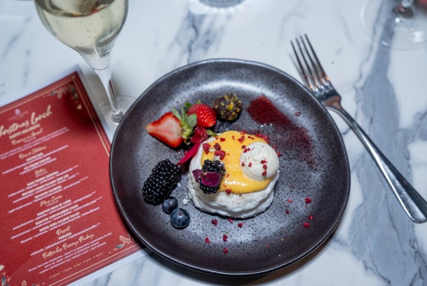 Looking down at a lemon and cream dessert with berry garnishes, a red menu and a glass of champagne. 