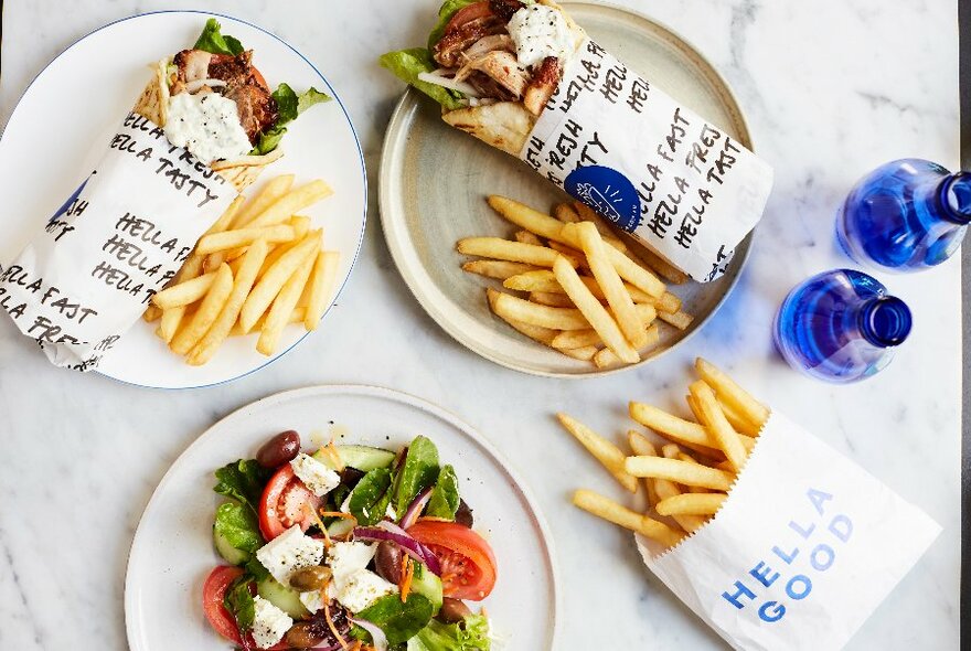 Souvlakis and chips wrapped in paper, a Greek salad and blue bottles on a marble table