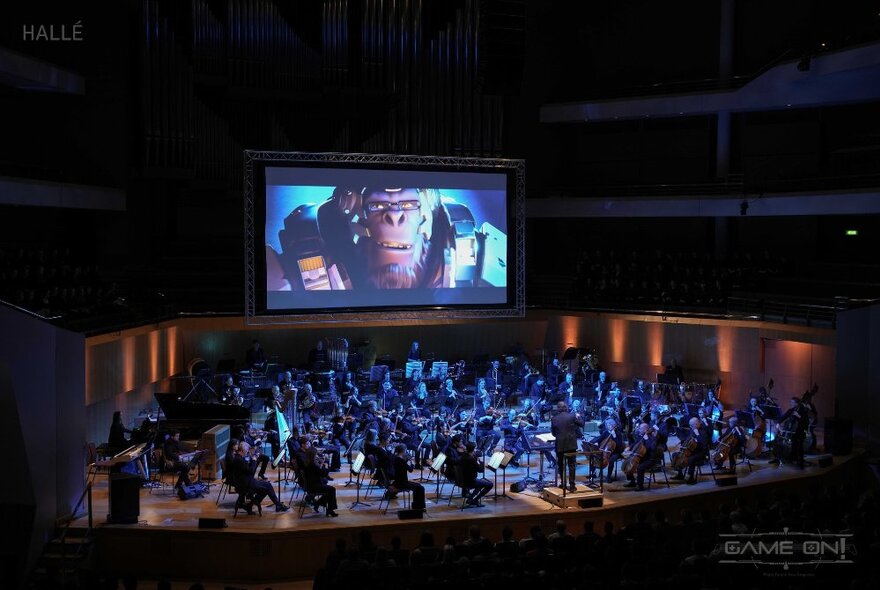 An orchestra performing live on stage while a movie plays on the screen behind.