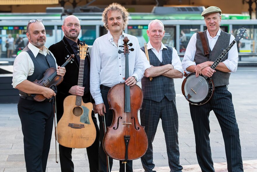 Portrait of gypsy-bluegrass ensemble, Blue Grassy Knoll, in front of a Melbourne tram stop, each band member holding their instrument.