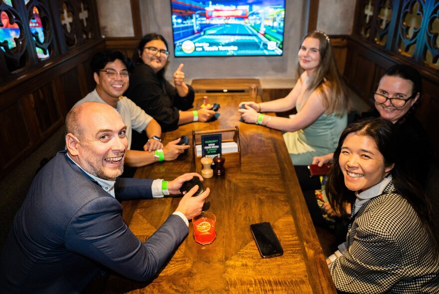 Gamers seated around a table holding consoles in front of a large screen.