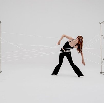 Still of a performance art installation featuring sculptures  and a human figure moving in the centre, white strings from their waist attached to the sculptures.