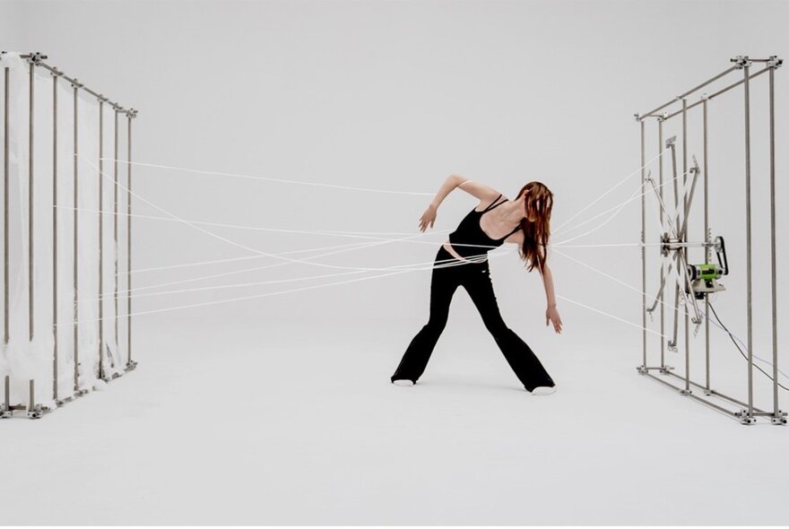 Still of a performance art installation featuring sculptures  and a human figure moving in the centre, white strings from their waist attached to the sculptures.