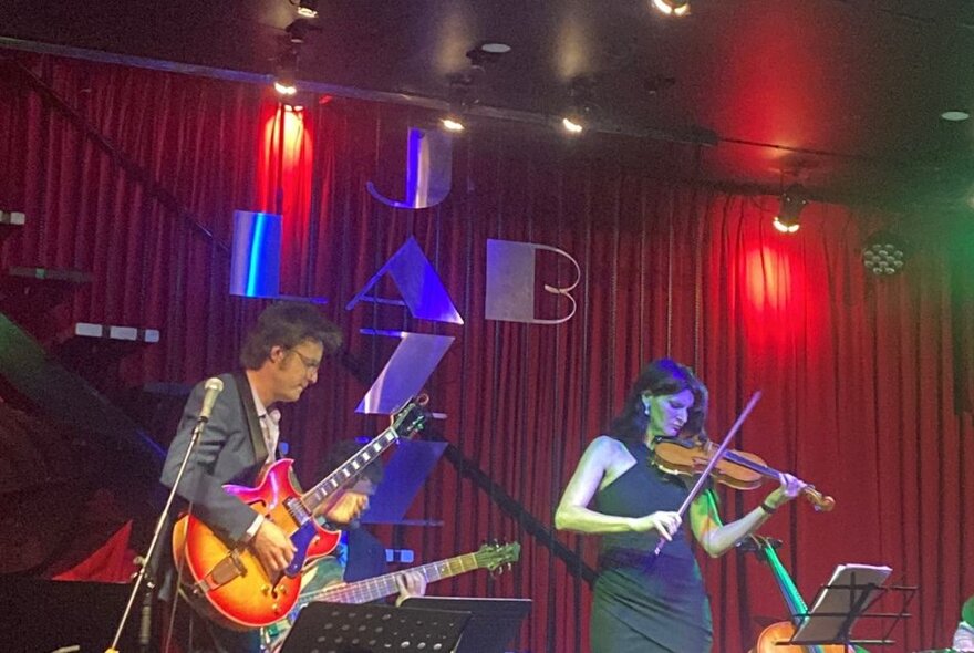 A guitarist and violinist performing live on stage, stage lights behind them shining on a red curtain.