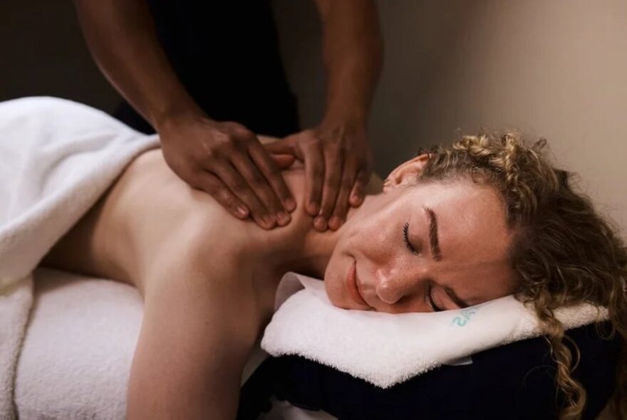 A woman with curly hair lying face down on a massage table, receiving a back massage from a therapist whose hands are kneading her upper shoulder.