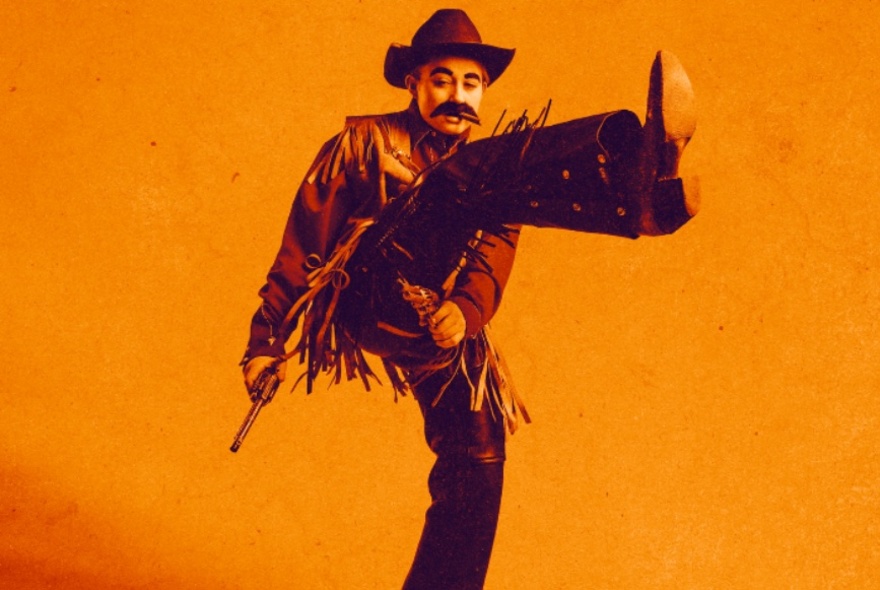 Comedian, Lil Wenker, dressed as a Wild West cowboy, posing with one leg up and gun in hand.