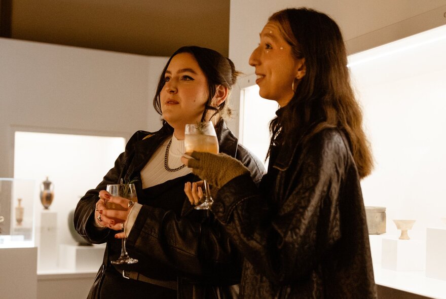 Two women in a gallery space looking at something with wine glasses in hand. 