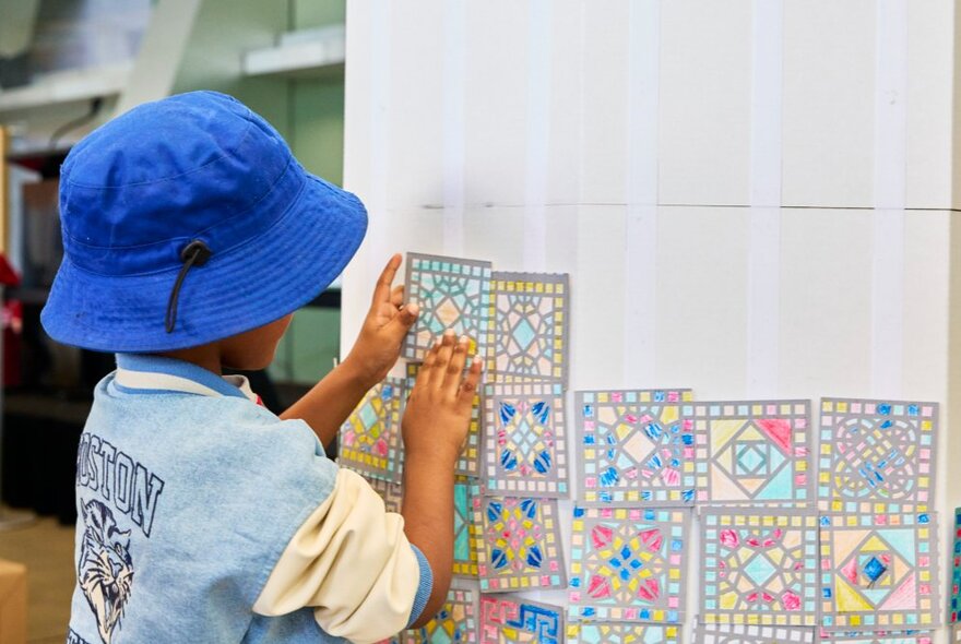 The back of a young child adding a hand-coloured small tile to a participatory art wall of tiles at a Melbourne Museum activity.