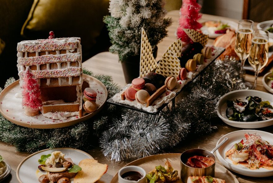 A table at a restaurant filled with plates of food, including a festive gingerbread house, and a  centrepiece of a tray of sweet treats resting on tinsel.