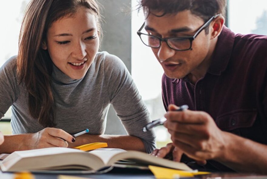 Two people smiling, sitting together and looking closely at an open book on a table.