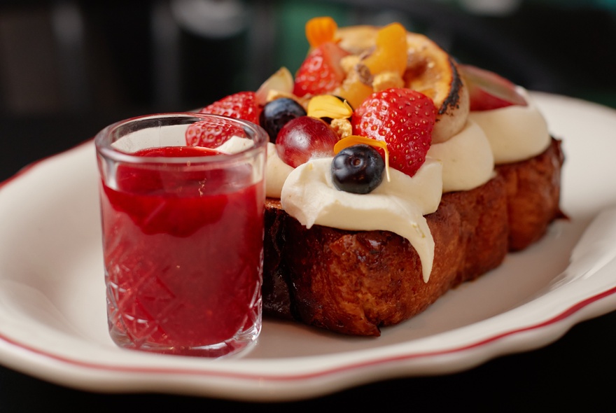 A slice of cake topped with cream and berries on a white plate with a side jar of red syrup. 