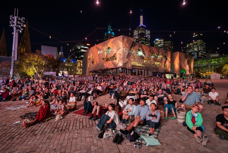 A crowd of people seated on deck chairs, beanbags or picnic blankets, watching an open air film screening at Melbourne's Fed Square.