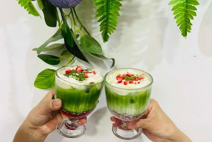 Two hands holding chilled matcha drinks in glasses close together.