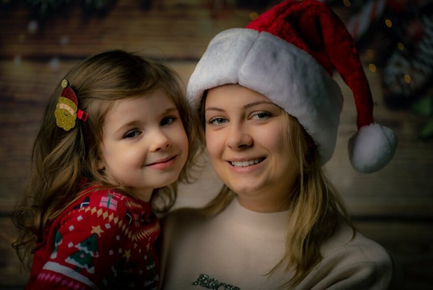 A woman holds a little girl up while wearing a Santa hat, both smiling. 