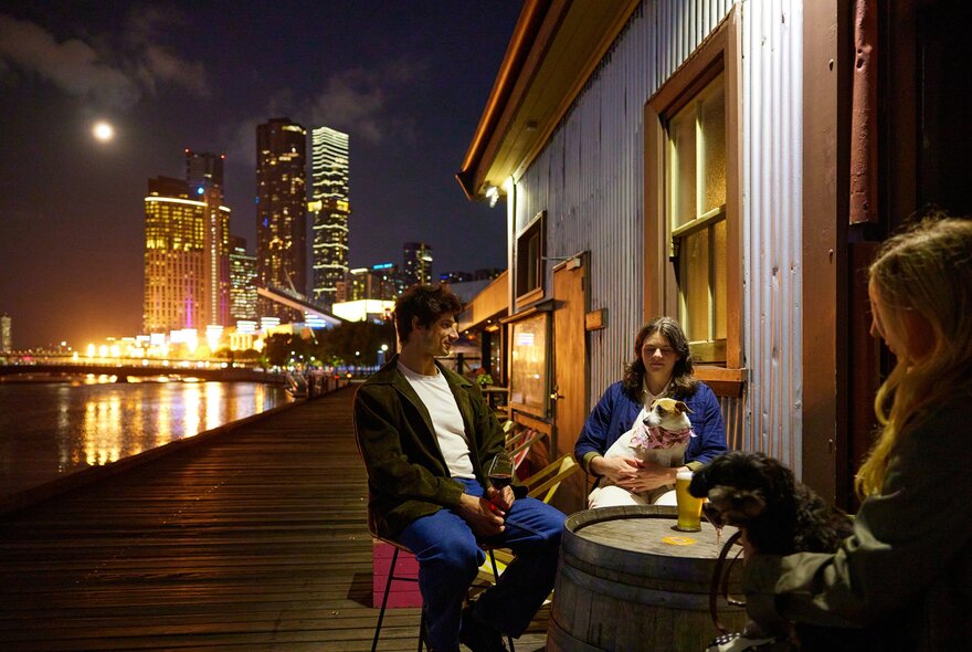 A group of friends and their dogs are drinking beers at a riverside bar there is a view of Melbourne's skyline and flame sculptures going off in the background.