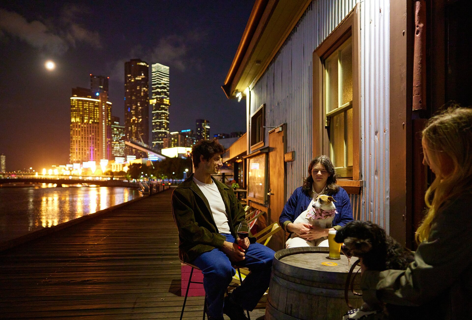 A group of friends and their dogs are drinking beers at a riverside bar there is a view of Melbourne's skyline and flame sculptures going off in the background.