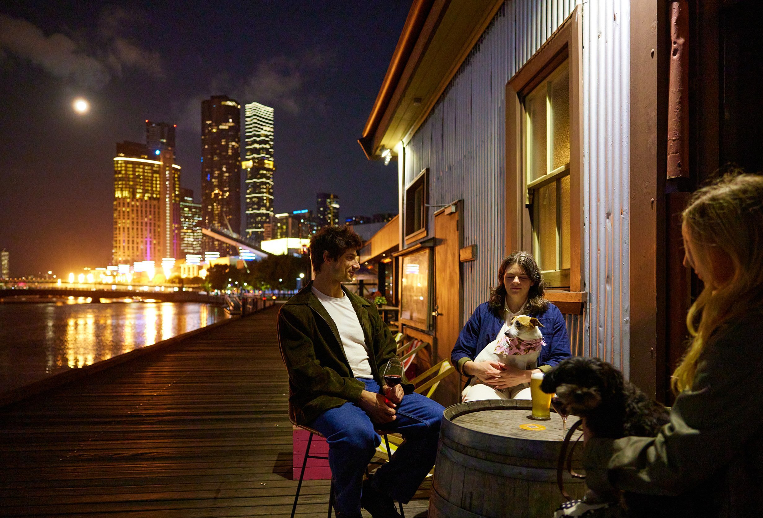 A group of friends and their dogs are drinking beers at a riverside bar there is a view of Melbourne's skyline and flame sculptures going off in the background.