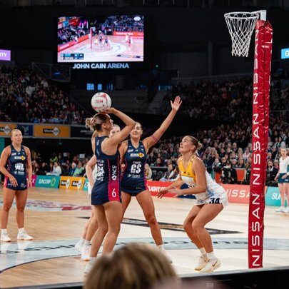Netball teams, the Melbourne Vixens and the Sunshine Coast Lightening, play netball in an indoor stadium with a large crowd.