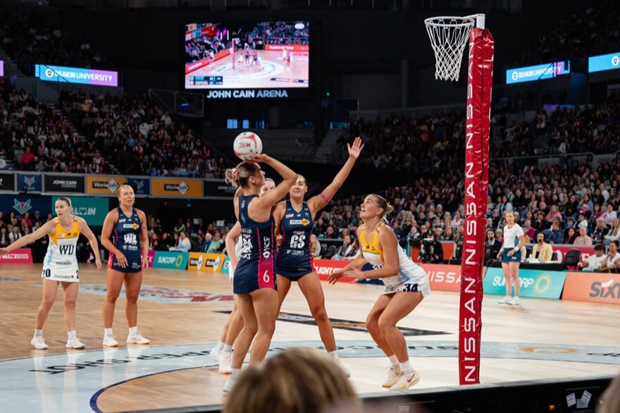 Netball teams, the Melbourne Vixens and the Sunshine Coast Lightening, play netball in an indoor stadium with a large crowd.