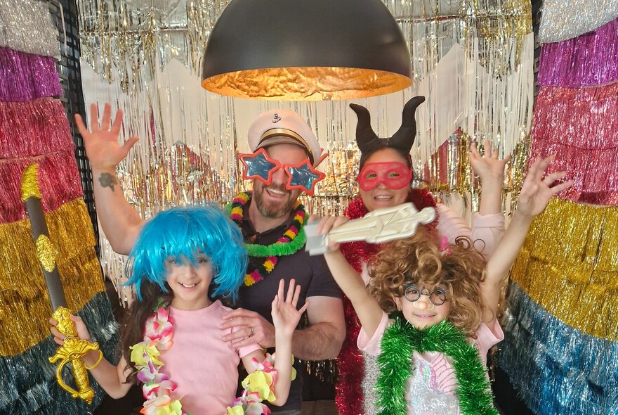 A family of four posing together in a large photo booth, wearing costumes, wigs and other props such as hats, masks, and glasses.