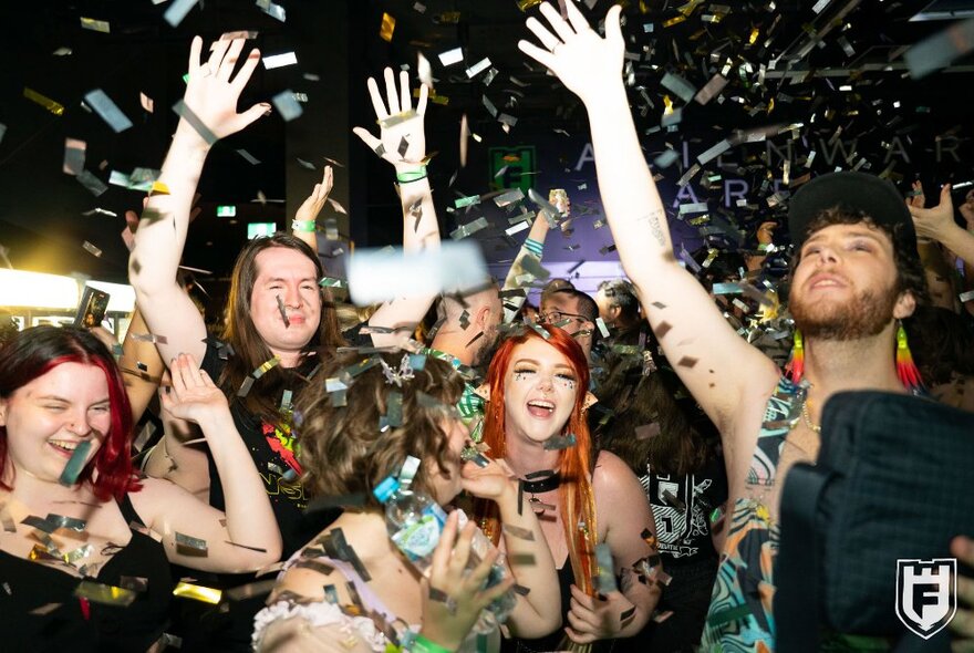 A crowd of people celebrating New Year's Eve with arms in the air and large gold confetti flying around.