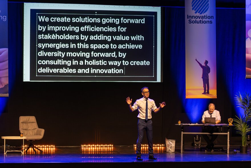 Performers on stage in front of a large powerpoint type display screen of office jargon. 