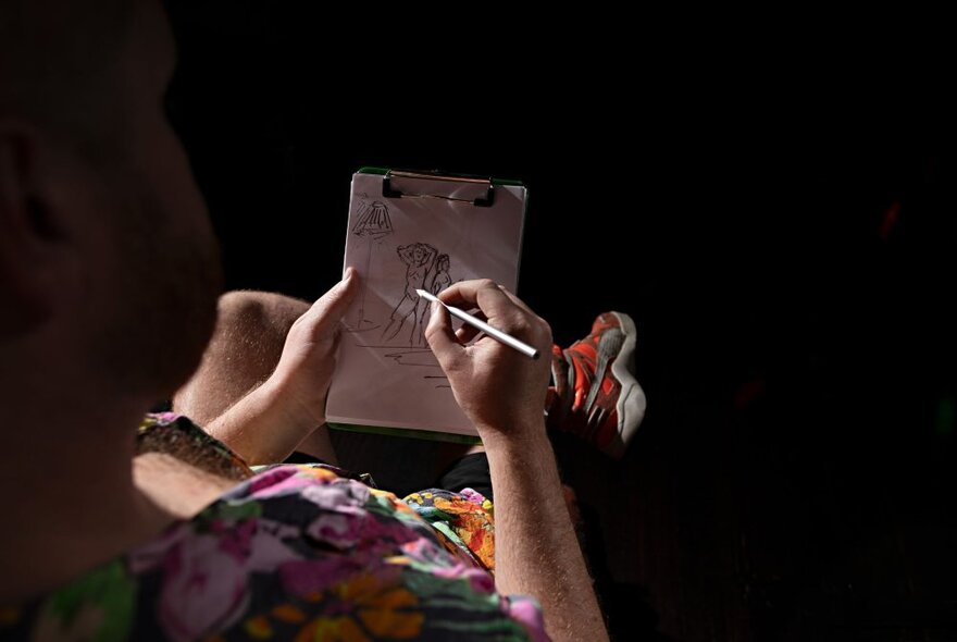 A person drawing with a clipboard and pencil, seen from over their shoulder.