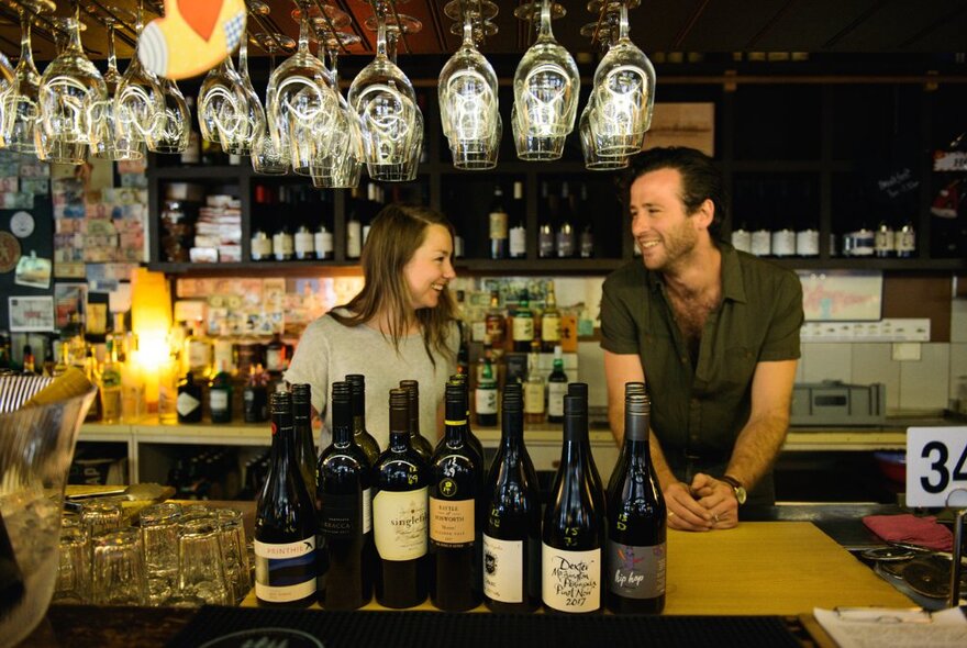 Two people smiling at each other at a bar with bottles in the front and glasses hanging upside down. 