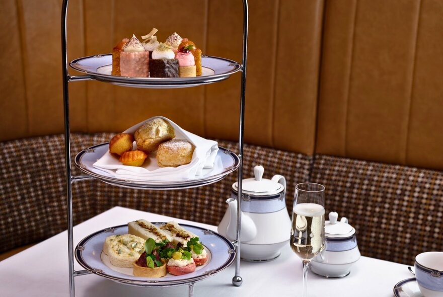 A three-tiered platter of savoury and sweet bites, a pot of tea and a glass of Champagne, on a dining table as part of a high tea menu.