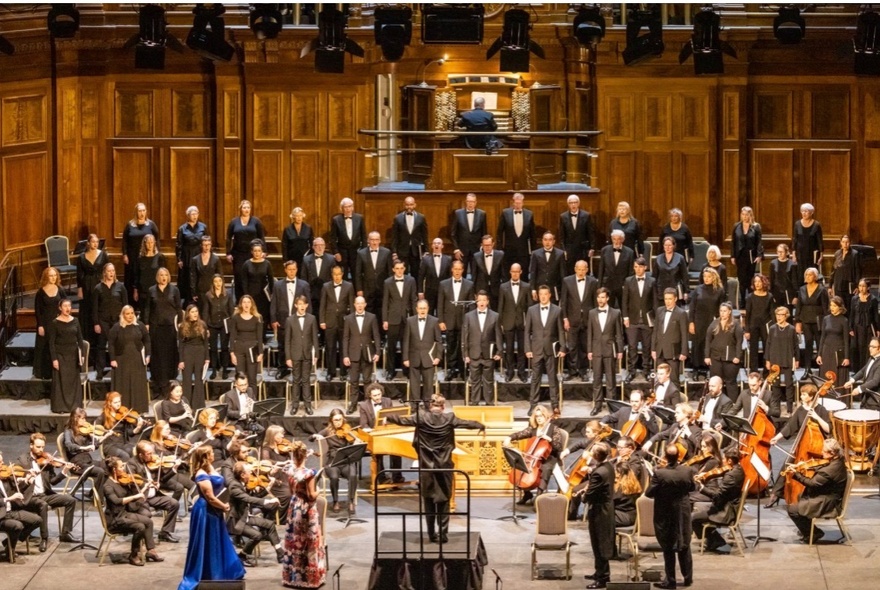 A large choir, the Royal Melbourne Philharmonic Choir standing on tiered rows behind an orchestra, performing at the historic Melbourne Town Hall.