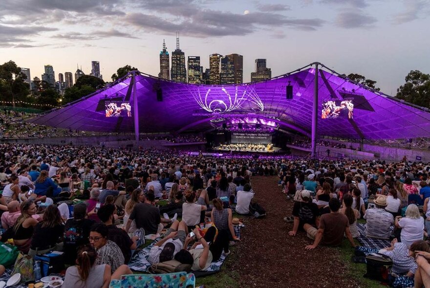 A large crowd seated on the lawns surrounding the Sidney Myer Music Bowl at dusk, with the venue in mid-distance bathed in purple light, an orchestra playing on the stage, and the outline of city buildings against the sky in the background.