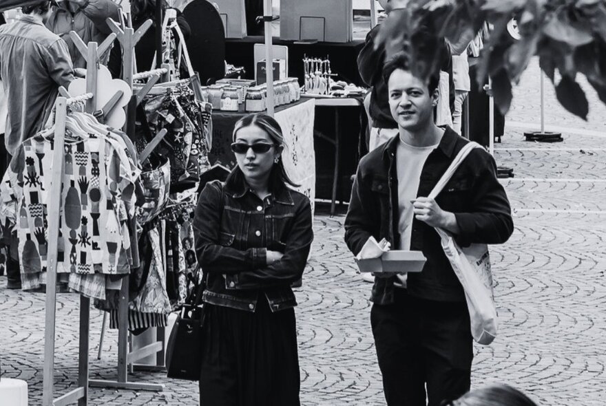 Black and white photograph of a young couple at an open air market, clothing racks in the background.