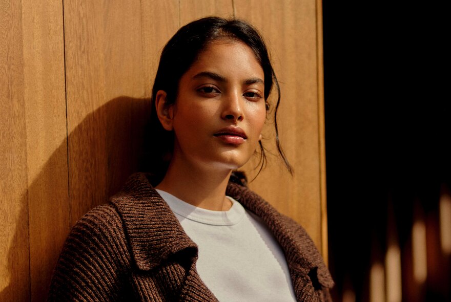 A female fashion model wearing Incu clothing, leaning against a wood panelled wall.
