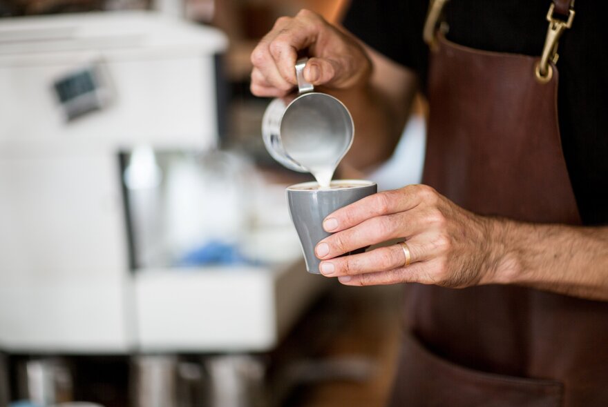 A barista wearing an apron and pouring milk from a jug into a cup of coffee.
