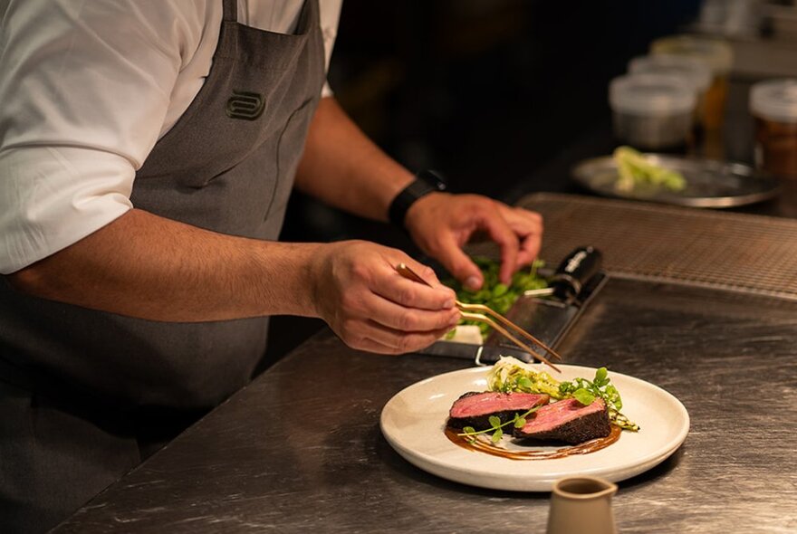 A chef in a kitchen putting the finishing garnishes on a plate of food that rests on a stainless steel bench.