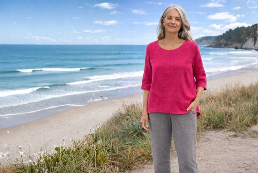 A woman modelling a casual pink top and grey slacks by the beach with waves rolling in and sand dunes visible. 
