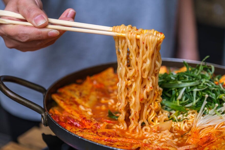 Hands using chopsticks to lift noodles from a iron dish of noodles in curry.