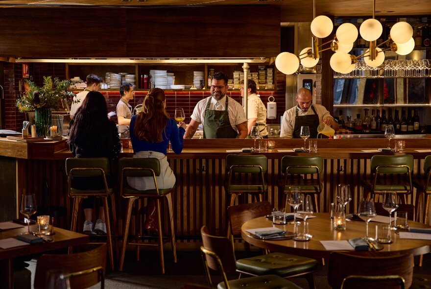 Two people seated at a high bar in a restaurant overlooking the chef's open kitchen and preparation area, with tables set for a meal service in the foreground.